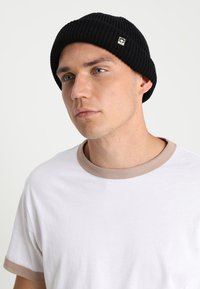 Svart ribbestrikket lue med en liten gulllogo, iført sammen med en hvit t-skjorte med en lys beige krage.