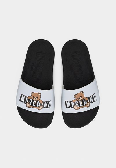 Ζευγάρι μαύρων σανδαλιών τύπου σλάιντ με λευκές λουρίδες που φέρουν ένα καφέ αρκουδάκι και τη λέξη «Moschino» με έντονα μαύρα γράμματα.