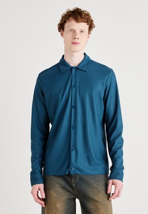 Teal long-sleeve shirt with a collar, featuring a button-up front and ribbed texture, paired with faded denim pants.