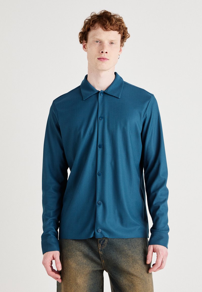 Teal long-sleeve shirt with a collar, featuring a button-up front and ribbed texture, paired with faded denim pants.