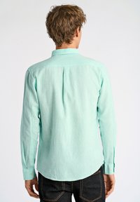 Jasnoniebieska koszula z długim rękawem, wykonana z fakturowanej tkaniny, z klasycznym kołnierzykiem i zaokrąglonym dołem, noszona na ciemnych dżinsach.