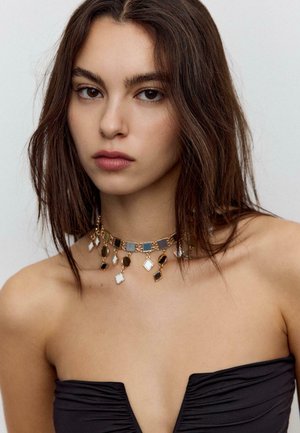 Nuori nainen, jolla on pitkät ruskeat hiukset. Hänellä on yllään olkaimeton musta paita ja kultainen kaulakoru, jossa on mustavalkoisia geometrisia riipuksia.