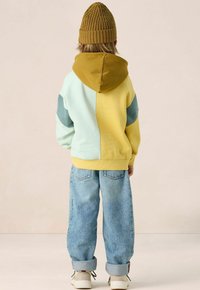 Kind dat met de rug naar de camera staat, gekleed in een colorblock hoodie in geel en lichtblauw, blauwe jeans, beige sneakers en een mosterdkleurige gebreide muts.