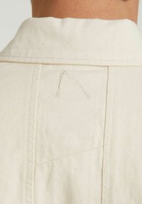 Camicia beige naturale con un tessuto testurizzato, colletto a punta e cuciture visibili con un distintivo dettaglio del filo sul retro.