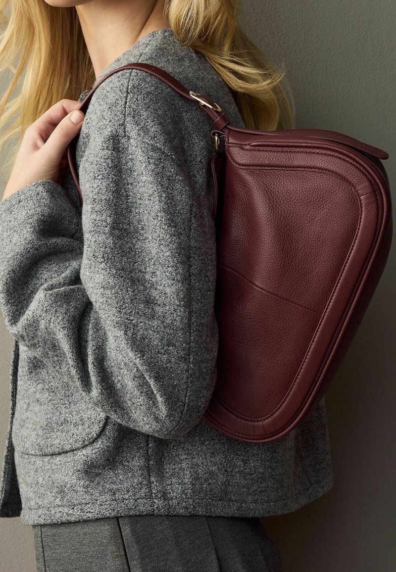 Femme portant une veste grise texturée et un sac à main en cuir rouge foncé sur l'épaule devant un fond uni.