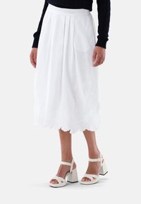 Gonna bianca, lunga fino al ginocchio, realizzata in tessuto leggero, con delicati ricami lungo l'orlo e pieghe verticali. Abbinata a sandali con tacco spesso.