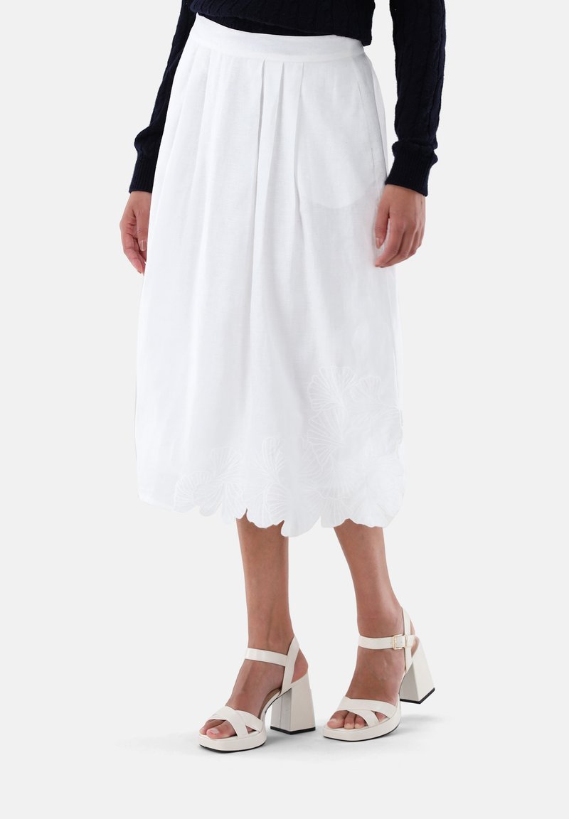 Gonna bianca, lunga fino al ginocchio, realizzata in tessuto leggero, con delicati ricami lungo l'orlo e pieghe verticali. Abbinata a sandali con tacco spesso.