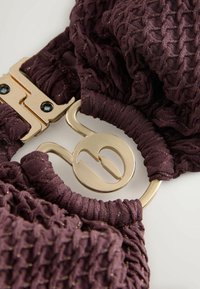 Fioletowa tkanina o fakturze ze złotymi drobinkami, z zapięciem w złotym odcieniu i charakterystycznymi ozdobami w kształcie logo.