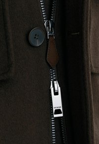 Detailní záběr tmavě hnědého vlněného kabátu s černým knoflíkem a stříbrným jezdcem zipu s vyrytým nápisem "REISS" a koženou záložkou na zipu.