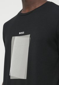 Čierne tričko z bavlny s šedým obdĺžnikom so svetlými zvislými čiarami a nad dizajnom nápisom „BOSS“.