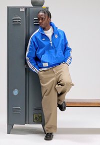 Blå lättviktsjacka med vita ränder, beige lösa byxor och svarta skor, lutad mot ett grönt skåp med en träbänk.