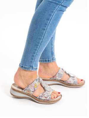Feet wearing light blue skinny jeans and silver open-toe sandals with decorative cutouts and a low wedge heel on a white background.