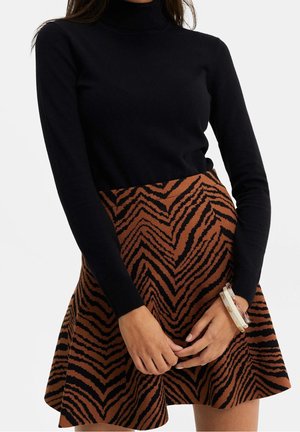 Mujer que lleva un suéter de cuello alto negro de manga larga y una falda marrón con rayas negras tipo cebra, con las manos entrelazadas y una pulsera blanca.