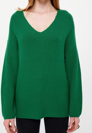Donna che indossa un maglione a coste verde con scollo a V e maniche lunghe, abbinato a pantaloni neri su uno sfondo semplice.