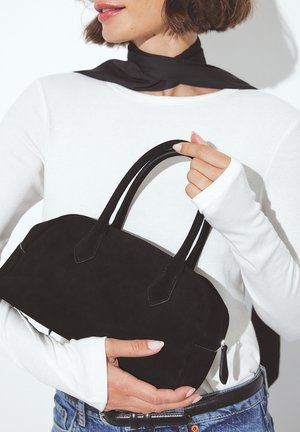 Borsa a tracolla in suede nero con due manici corti, forma arrotondata, chiusura zip e dettagli di cucitura sottili. Il modello indossa una maglietta bianca a maniche lunghe.