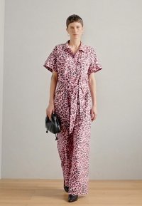 Rózsaszín leopárdmintás overál, rövid ujjakkal és megkötős derékkal, könnyű anyagból készítve. A modell egy fekete táskát tart a kezében.