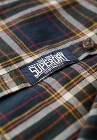 Camicia di flanella con motivo a quadri in blu scuro, bianco e rosso con accenti gialli; include un'etichetta blu con la scritta "Superdry" e dettagli con bottoni marroni.
