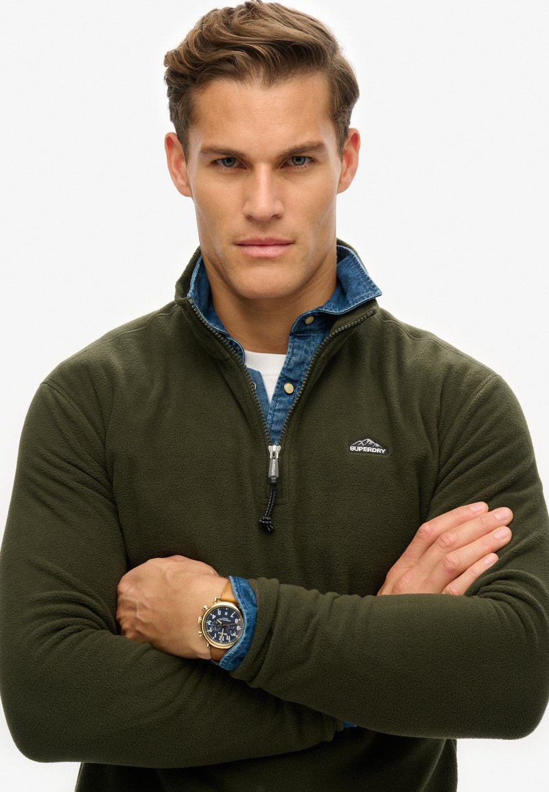 Donkergroen fleece jack met kwart-rits en een marineblauwe denim kraag, met een zwart logo op de borst en een ritssluiting met trekkuip. Gouden horloge om de pols.