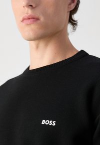 Czarna dzianinowa bluza z okrągłym dekoltem i ściągaczami, z białym logo "BOSS" na piersi.