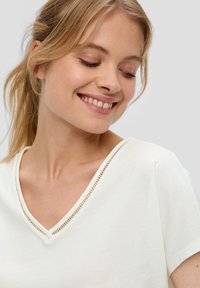 Biała bluzka z dekoltem w serek, z teksturowanym wykończeniem, krótkimi rękawami i miękkiego materiału. Projekt charakteryzuje się luźnym krojem i subtelnymi detalami przy dekolcie.