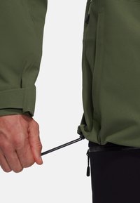 Main tirant sur le cordon noir pour resserrer l'ourlet d'une veste vert olive portée par-dessus un pantalon noir.