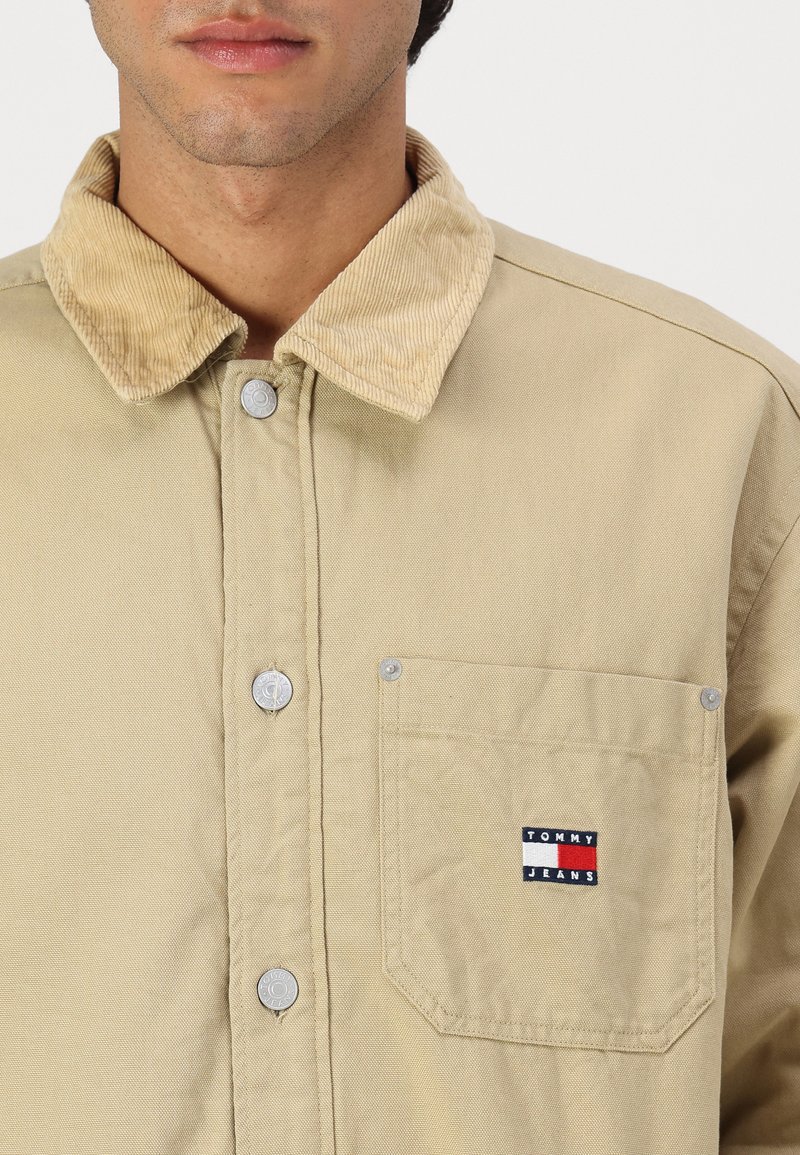 Chaqueta de camisa color beige con un cuello de pana, botones plateados, bolsillo en el pecho con un logo que presenta acentos en rojo, blanco y azul.