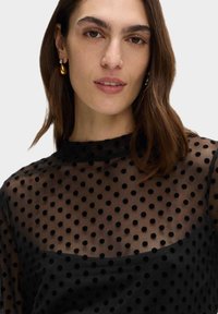 Svart prickig genomskinlig blus med hög krage, med texturerade prickar, hel långa ärmar och en detalj vid axelsömmen.