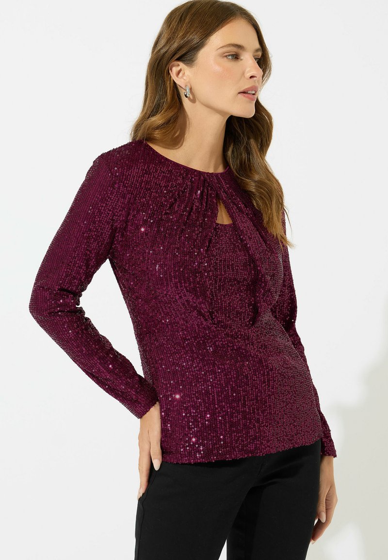 Top bordeaux a maniche lunghe con scollo arricciato e dettaglio a chiave, coperto di paillettes per un effetto testurizzato. Abbinato a pantaloni neri.