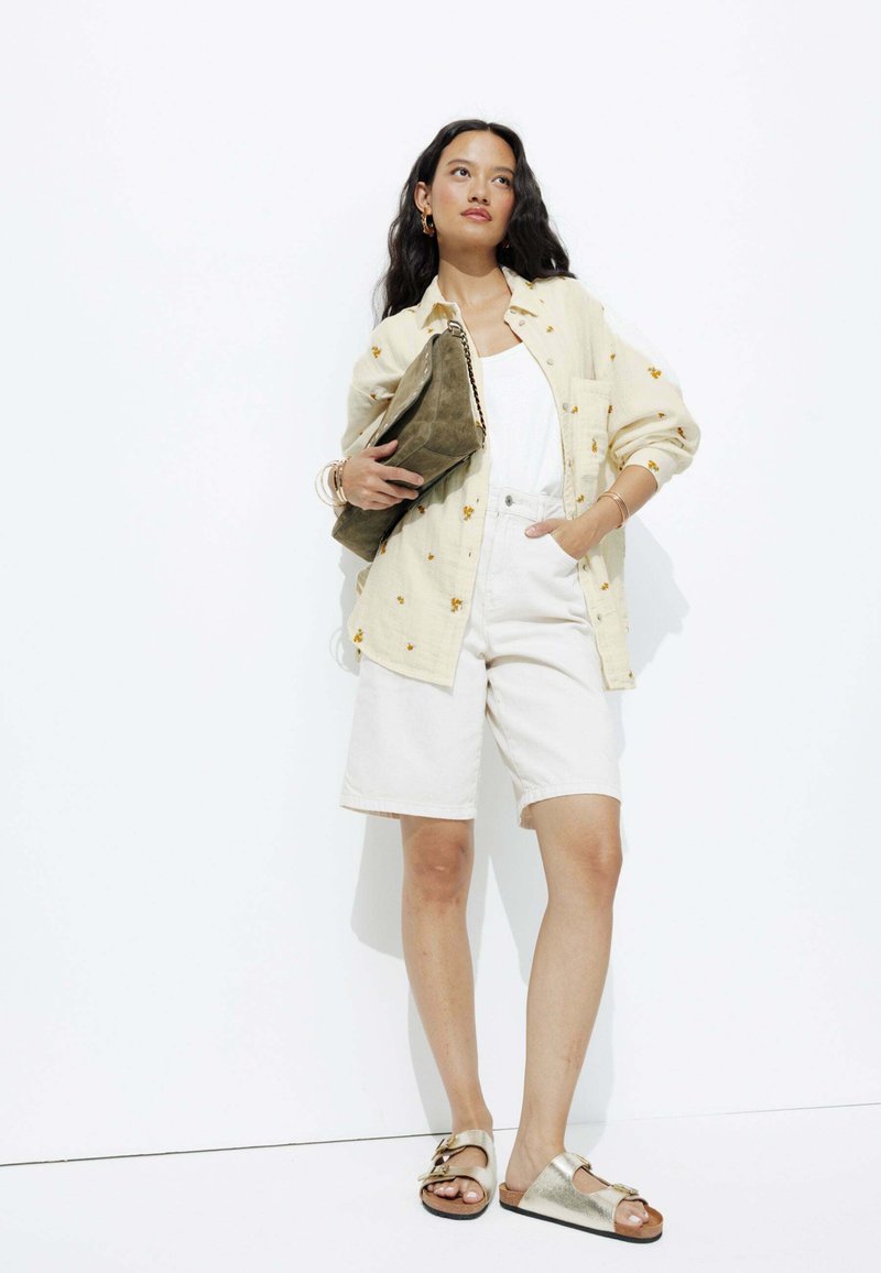 Chemise à boutons beige à motif floral, débardeur blanc, shorts clairs, sandales métalliques et tenant une pochette en cuir verte.
