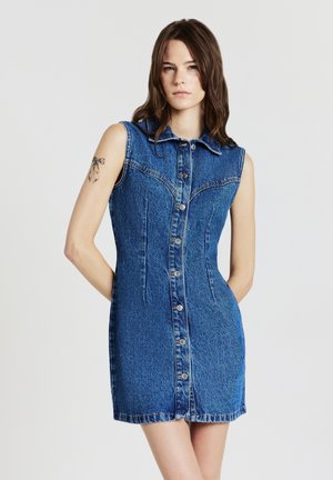 Junge Frau in einem ärmellosen, knöpfbaren blauen Jeanskleid steht mit Händen hinter dem Rücken vor einem weißen Hintergrund.