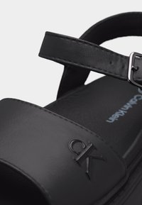 Μαύρο δερμάτινο σανδάλι με ρυθμιζόμενο λουράκι στον αστράγαλο και ανάγλυφο λογότυπο «CK» στο μπροστινό λουράκι, με εμφανές τμήμα της σόλας.