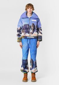 Chaqueta acolchada en azul y morado con estampado de paisaje montañoso; pantalones azules a juego con diseño similar; botas marrones; tela con textura.