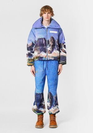 Chaqueta acolchada en azul y morado con estampado de paisaje montañoso; pantalones azules a juego con diseño similar; botas marrones; tela con textura.