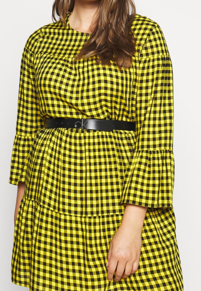Femme portant une robe à carreaux jaune et noire avec des manches évasées et une ceinture noire avec une boucle métallique "CK".