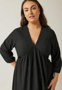 Schwarzes Kleid mit strukturiertem, plissiertem Design. Verfügt über einen tiefen V-Ausschnitt und drei Viertel lange Ärmel, mit einer lockeren Silhouette in der Taille.