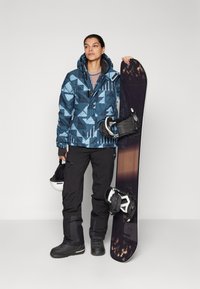 Giacca da sci blu con motivo geometrico, pantaloni da sci neri, stivali neri; tiene in mano uno snowboard nero e un casco bianco, con uno sfondo testurizzato.
