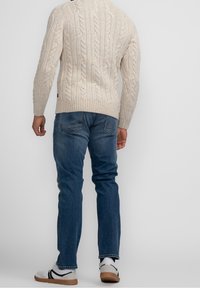 Homme portant un pull en maille torsadée crème, un jean bleu, des baskets blanches, debout de dos sur un fond gris clair uni.
