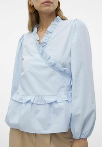 Blusa avvolgente azzurro chiaro con scollatura e orlo volant, maniche lunghe a palloncino e chiusura con fiocco laterale. Realizzata in tessuto morbido.