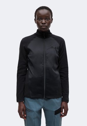 Personne portant une veste noire à fermeture éclair et un pantalon bleu, debout face à un fond clair uni.