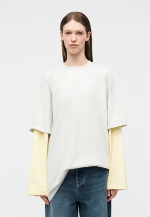 Γυναίκα με ίσια καστανά μαλλιά φοράει στρωτά, φαρδιά λευκά και ανοιχτόκίτρινα μακρυμάνικα πουκάμισα με μπλε τζιν, μπροστά σε μονόχρωμο φόντο.