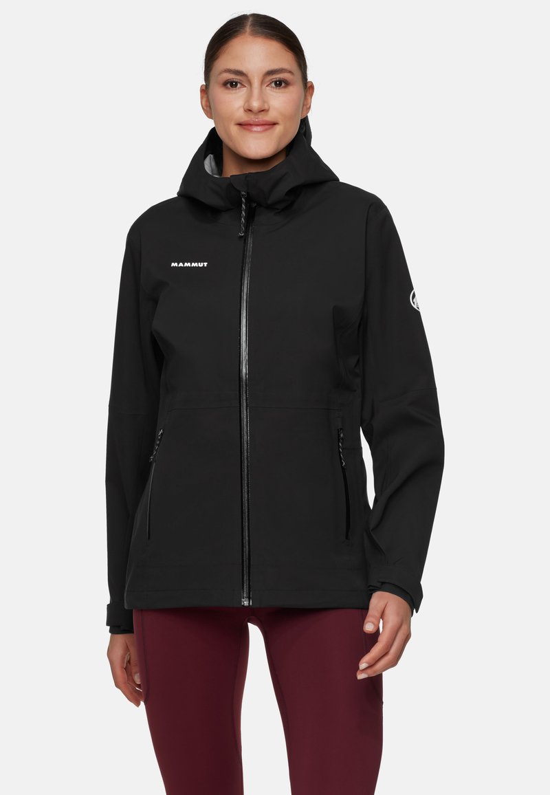 Kvinna i svart zip-up huva jacka med Mammut-logotyp och vinröda leggings, stående mot en enkel vit bakgrund.