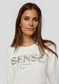 Junge Frau mit langen braunen Haaren, die ein weißes Oberteil trägt, auf dem vorne "SENSE Sensibility New" in metallischen Buchstaben aufgedruckt ist.