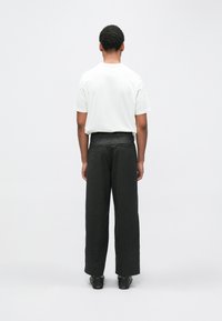 Balta īss piedurkņu T-krekls un tumši pelēkas, platas bikses ar smalku rakstu. Melni apavi noslēdz apģērbu. Skats no aizmugures.