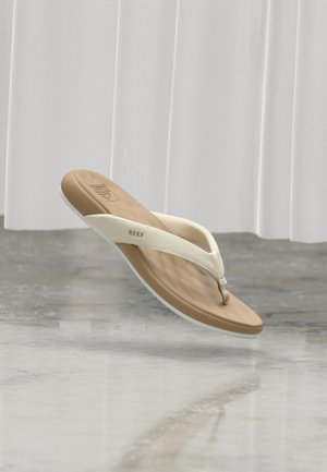 Una única sandalia flip-flop de la marca Reef en tonos marrón claro y blanco flotando sobre un suelo gris brillante con cortinas blancas de fondo.