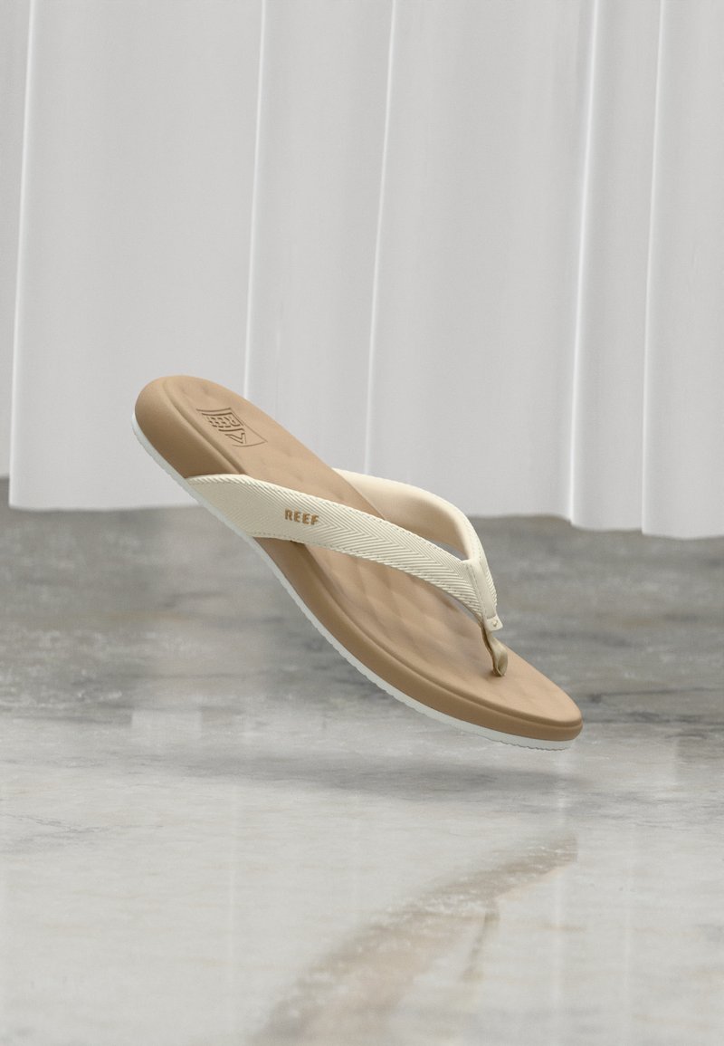 Une sandale tongs unique de la marque Reef, couleur marron clair et blanc, flottant au-dessus d’un sol gris brillant avec des rideaux blancs en arrière-plan.