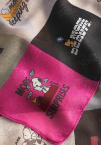 Zijde sjaal met een kleurrijk patchworkontwerp. Een opvallende roze vierkant met cartoonfiguren en de tekst "MEER VERRASSINGEN" omlijnd in zwart.