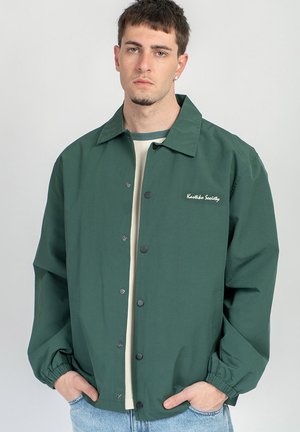 Jonge man die een donkergroene jas met knopen draagt, met "Koetko Society" geborduurd op de borst, handen in de zakken, neutrale achtergrond.