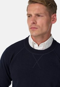 Navy mėlynas megztinis su apvaliu kaklu, raukšlėtomis rankovėmis ir tekstūruotu paviršiumi. Dėvimas virš baltos marškinių su apykakle, kuriame yra subtilių siūlių detalių.