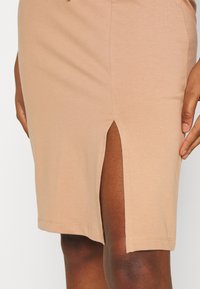 Gros plan sur une personne portant une jupe beige jusqu'aux genoux avec une fente frontale, montrant un teint de peau foncé et des mains reposant sur les côtés.
