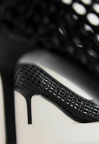 Musta kudottu korkokenkä, joka sisältää kärjeltään terävän muodon ja hoikan stiletto-korkean kannan, esittelee monimutkaista tekstuuria ja avointa muotoilua.